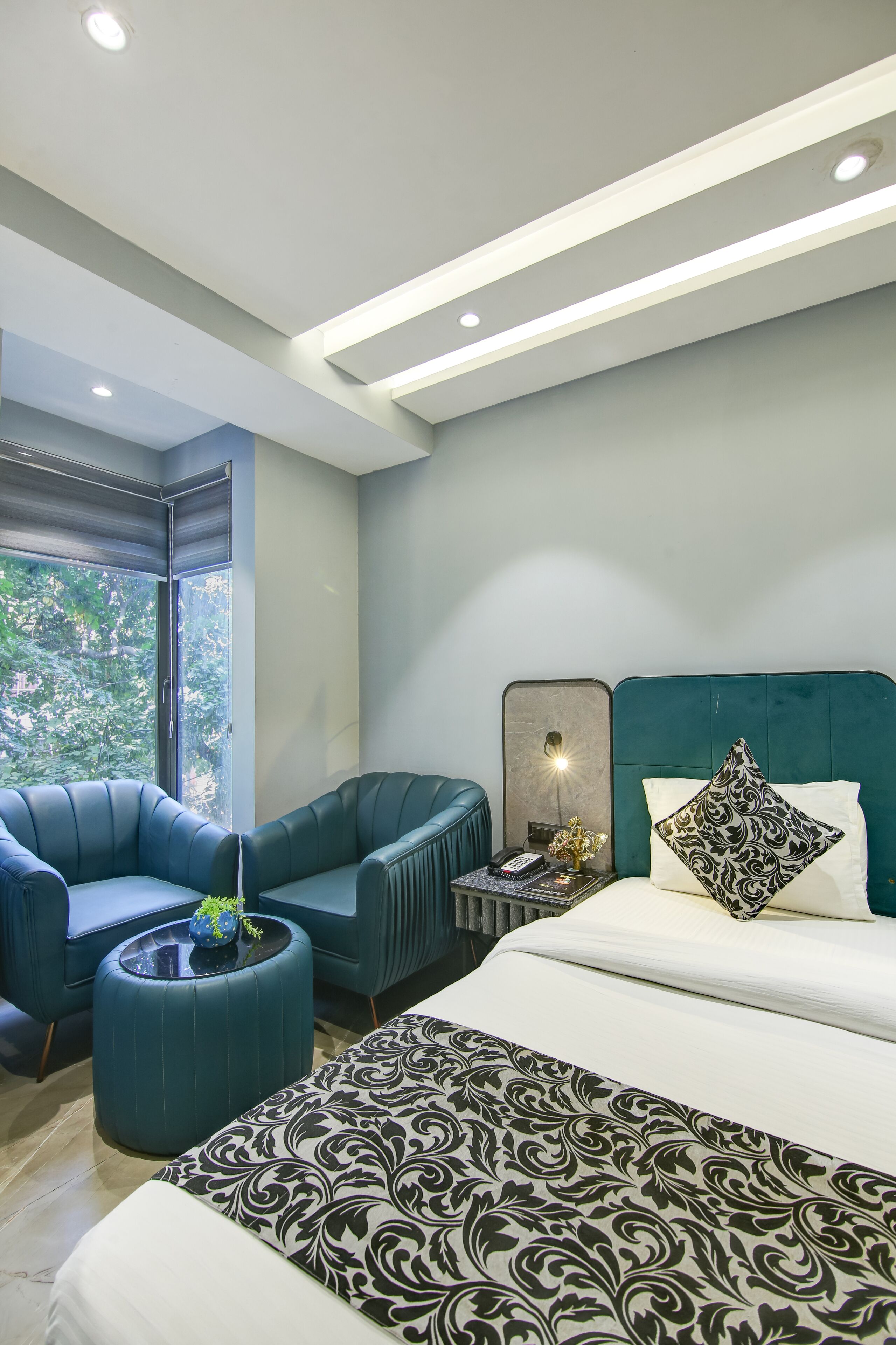 A modern bedroom with a large bed, a seating area with a chair and ottoman, a bedside table with a lamp, and a view of greenery through the window.