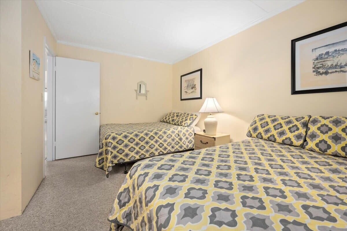 A bedroom with two beds, a nightstand, a lamp, a door, and a framed picture on the wall.