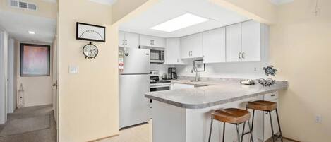 A modern kitchen with white cabinetry, a central island with stools, a microwave, and a clock on the wall.