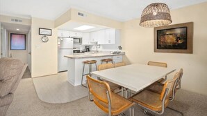 Een moderne keuken met witte kasten, een centraal eiland en een eethoek met een witte tafel en rieten stoelen.