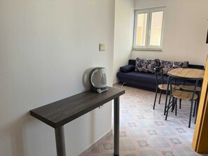 A small, modern room with a dark wooden table, a kettle, a sofa with patterned cushions, a round wooden table, and a window with blinds.