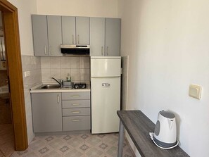 Eine kleine Küche mit grauen Schränken, einem weißen Kühlschrank, einer Spüle und einem Wasserkocher auf einer Arbeitsplatte.