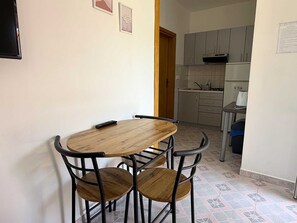 A dining area with a wooden table and four chairs, a kitchen in the background, and a door leading to another room.