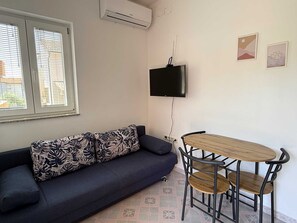 A room with a sofa, a TV mounted on the wall, a small table with chairs, and a window with blinds.