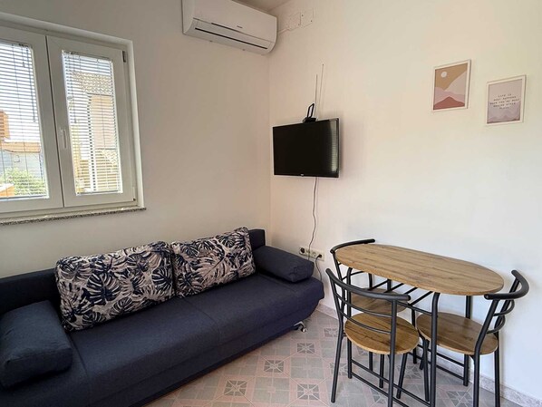 Ein Zimmer mit einer Couch, einem an der Wand montierten Fernseher, einem kleinen Tisch mit Stühlen und einem Fenster mit Jalousien.