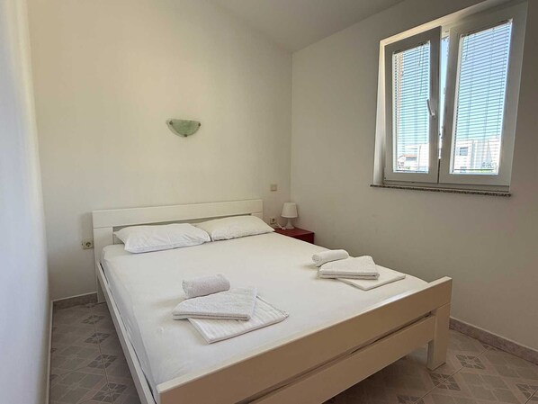 A single bed with white bedding and towels, a bedside table with a lamp, and a window with blinds.