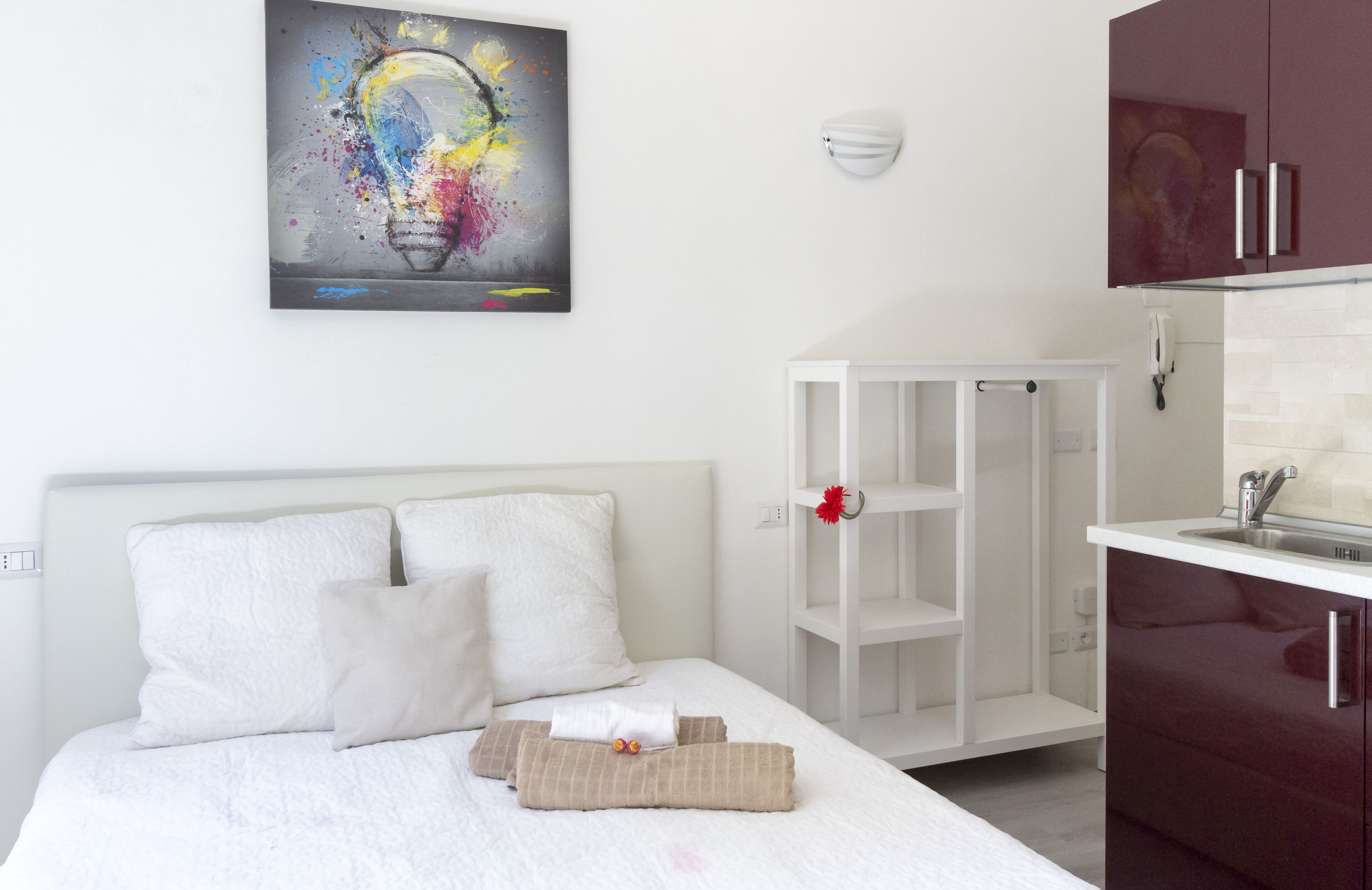 Een hotelkamer met een bed, witte kussens, een nachtkastje, een rode bloem, een witte kast en een kleurrijk abstract schilderij aan de muur.