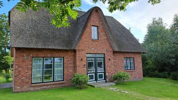 A brick house with a steeply pitched thatched roof, large windows, and a well-maintained lawn.