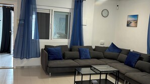 Una sala de estar moderna con un sofá seccional, una mesa de centro de vidrio y un ventanal con cortinas azules.