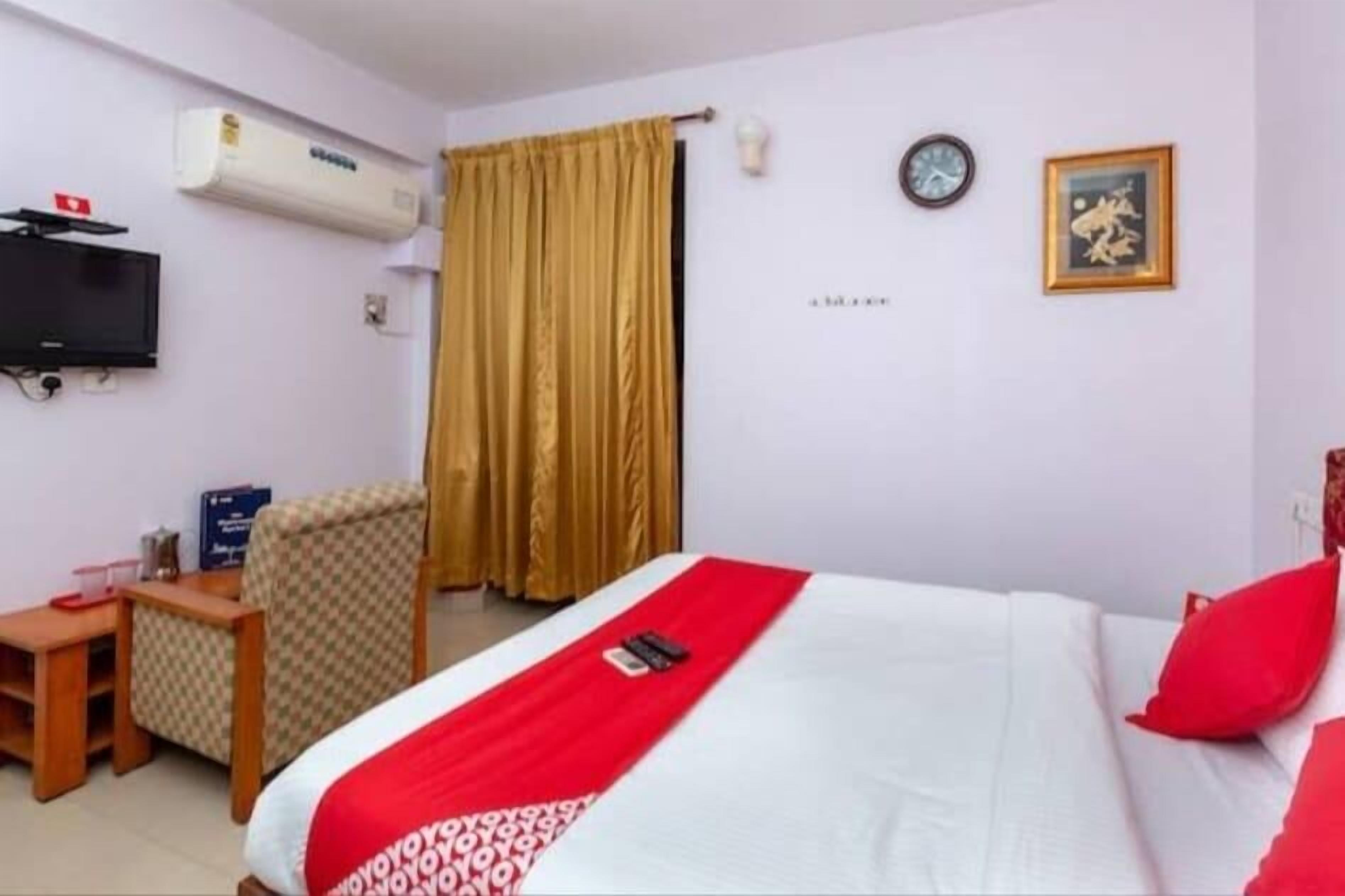 A hotel room with a bed, a television, an air conditioner, a desk, a chair, a clock, and a framed picture on the wall.