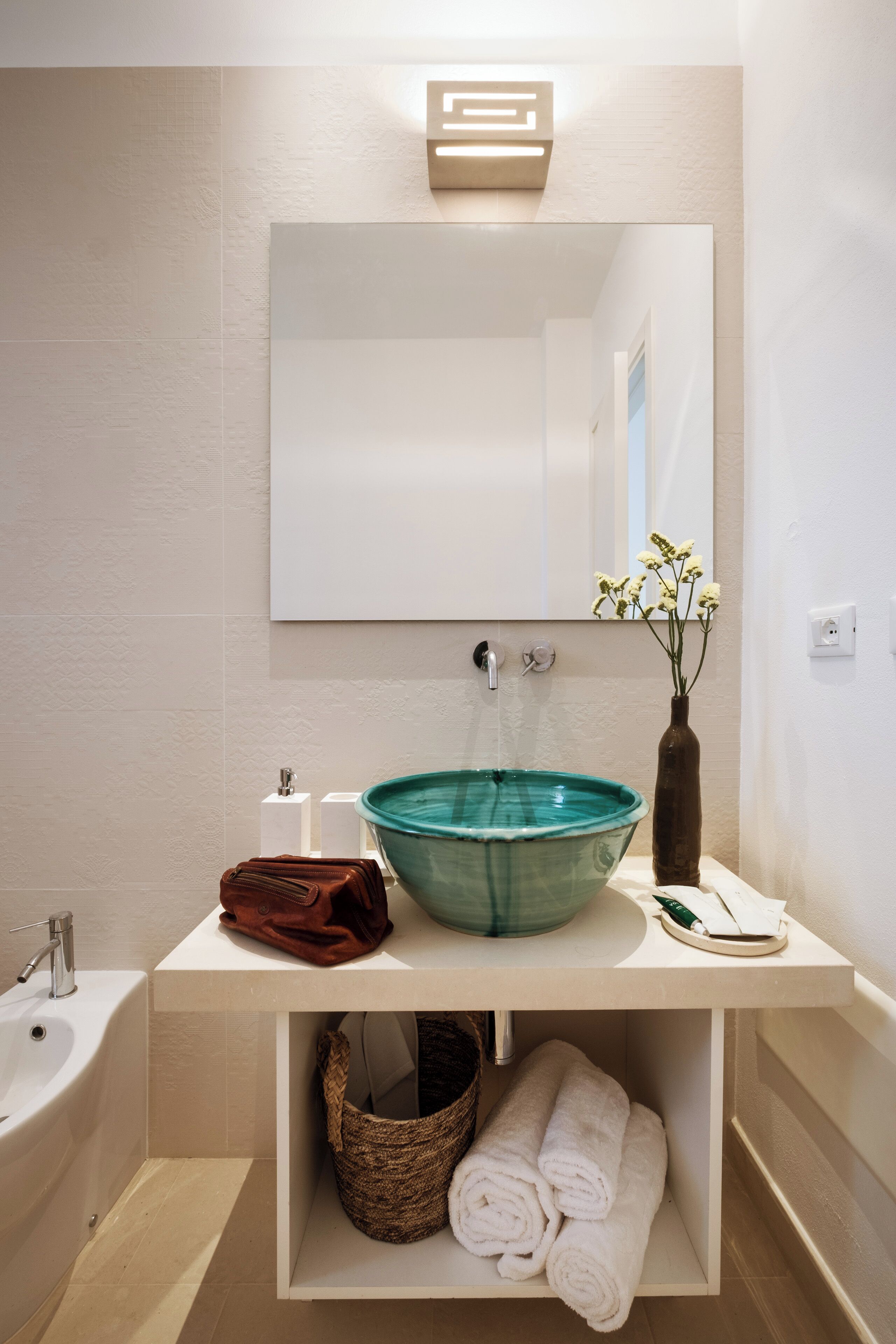 Un baño con un espejo grande, un lavabo de color verde, un jarrón marrón con flores, una toalla blanca y una cesta de mimbre.