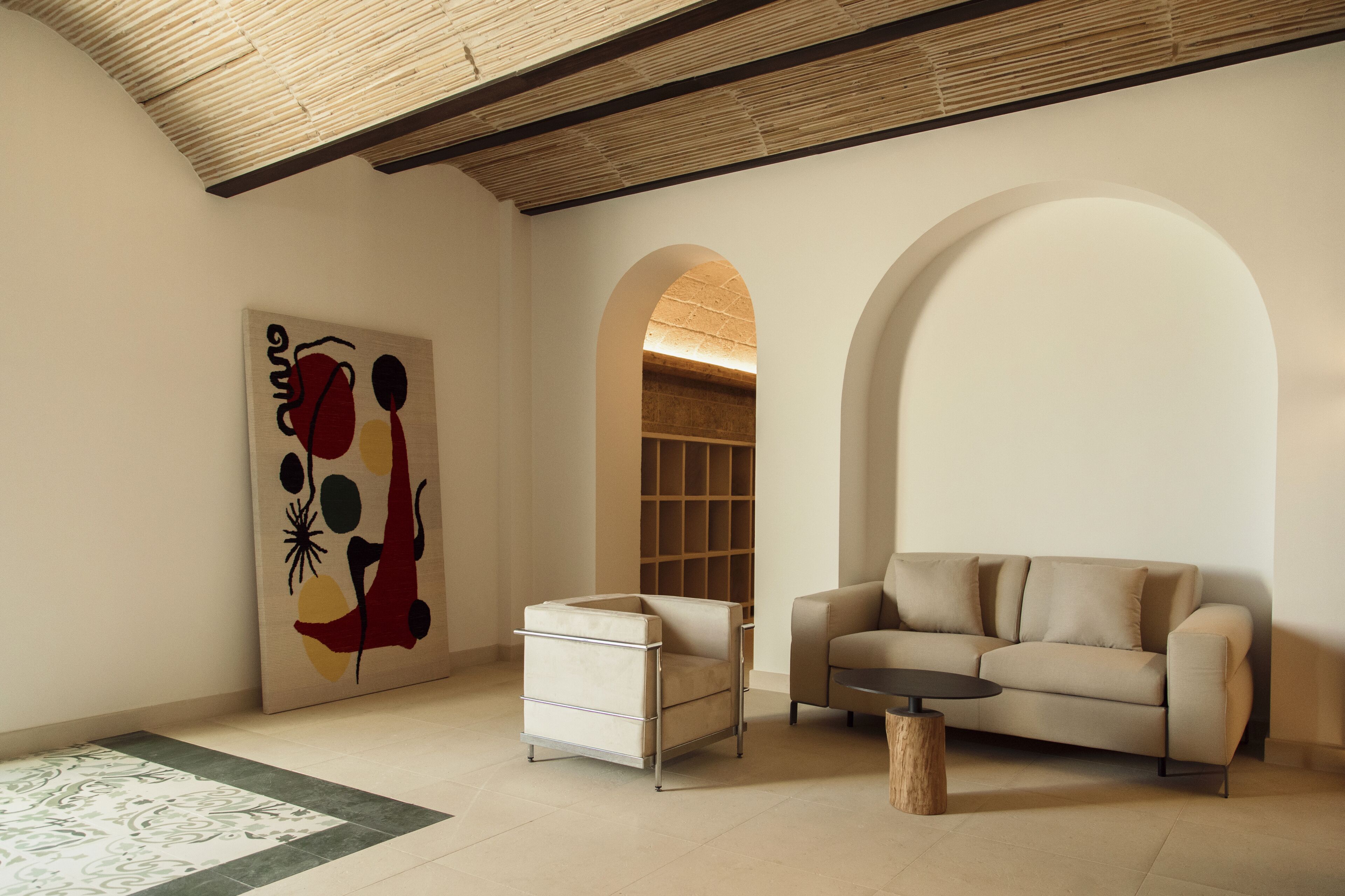 Una sala de estar moderna con un sofá, una mesita y una silla. Hay un cuadro en la pared y un arco que conduce a otra habitación.