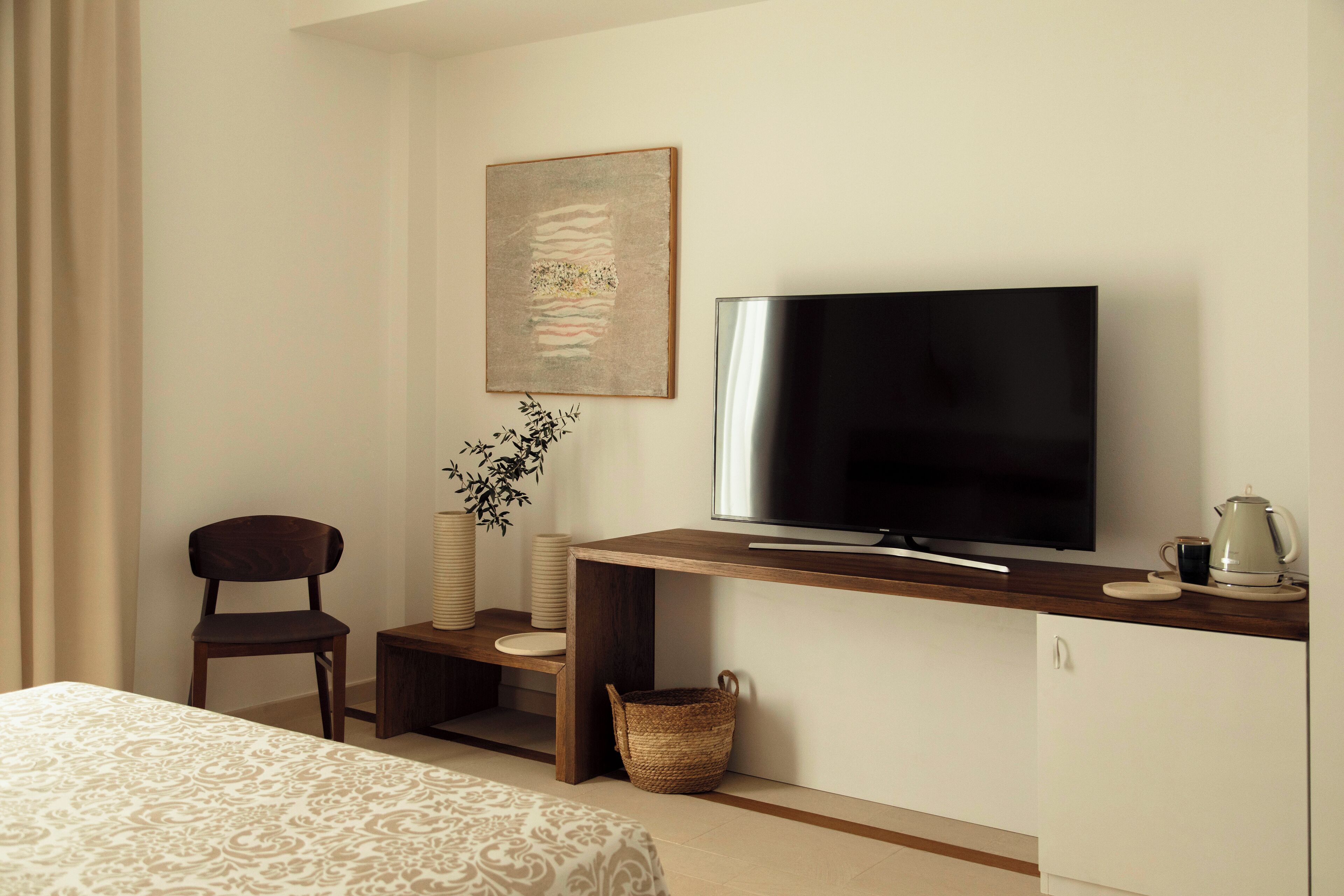 Una habitación de hotel con cama, televisor sobre un soporte de madera, una silla, una mesita, un adorno en la pared y una cesta.