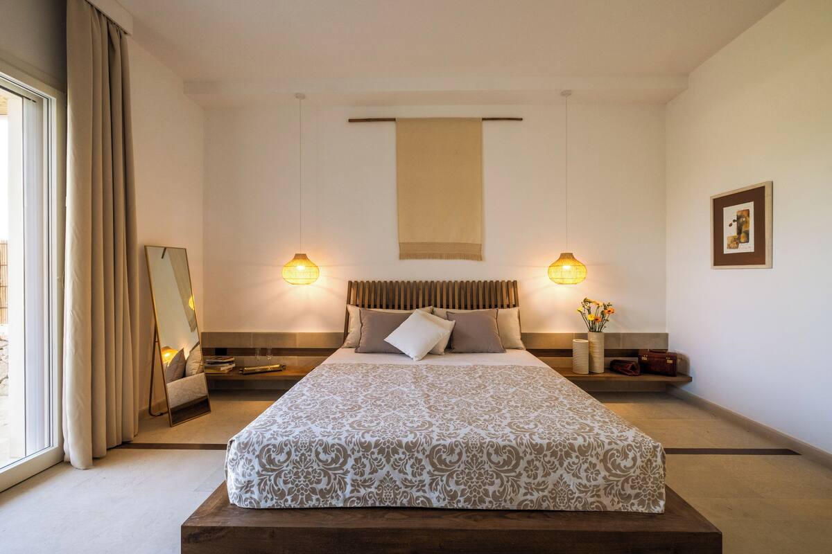 A modern hotel room with a large bed, two bedside tables, a nightstand, a large window with curtains, and a wall-mounted towel rack.
