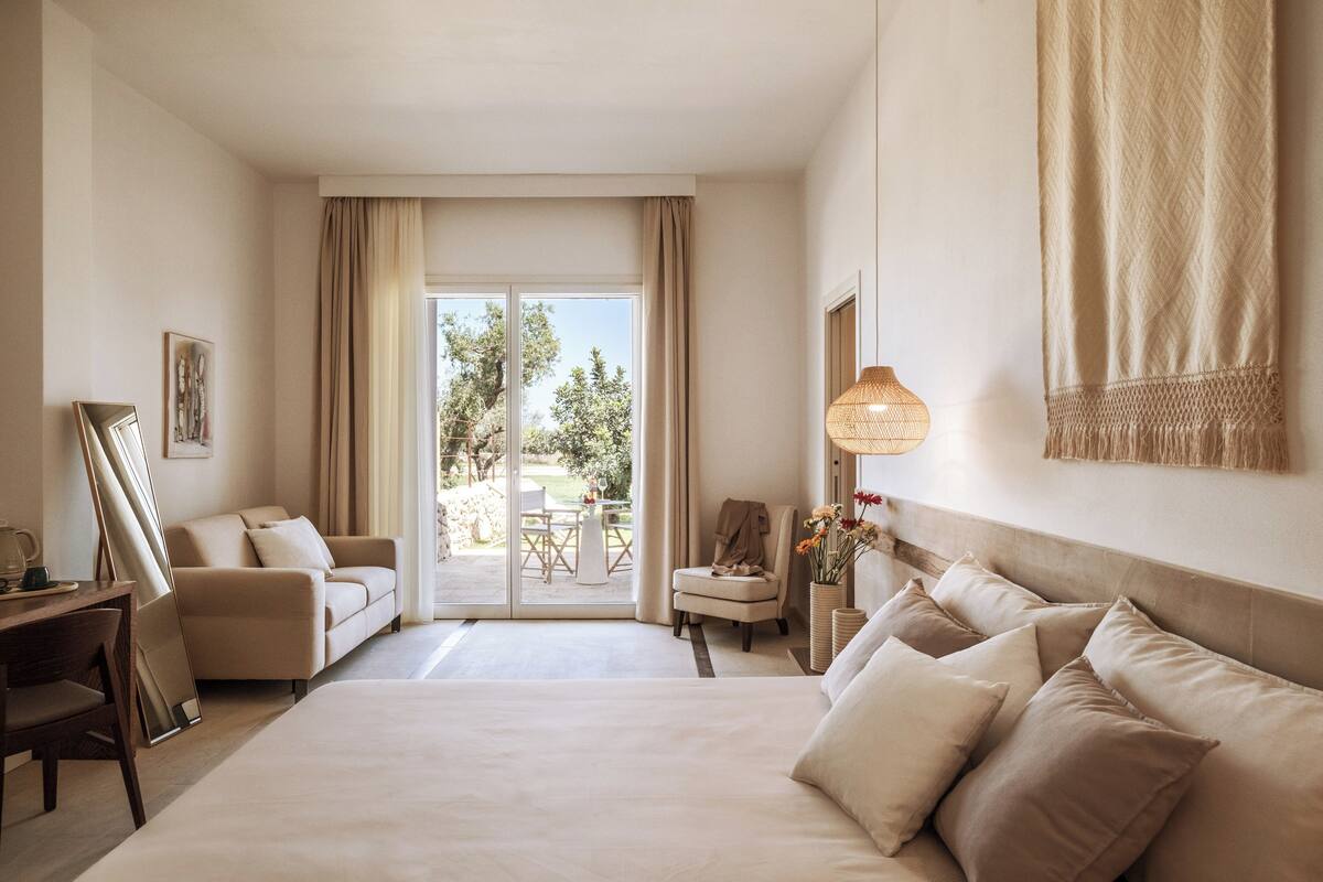 A hotel room with a large bed, a desk, a chair, and a view of the outdoors through a glass door.