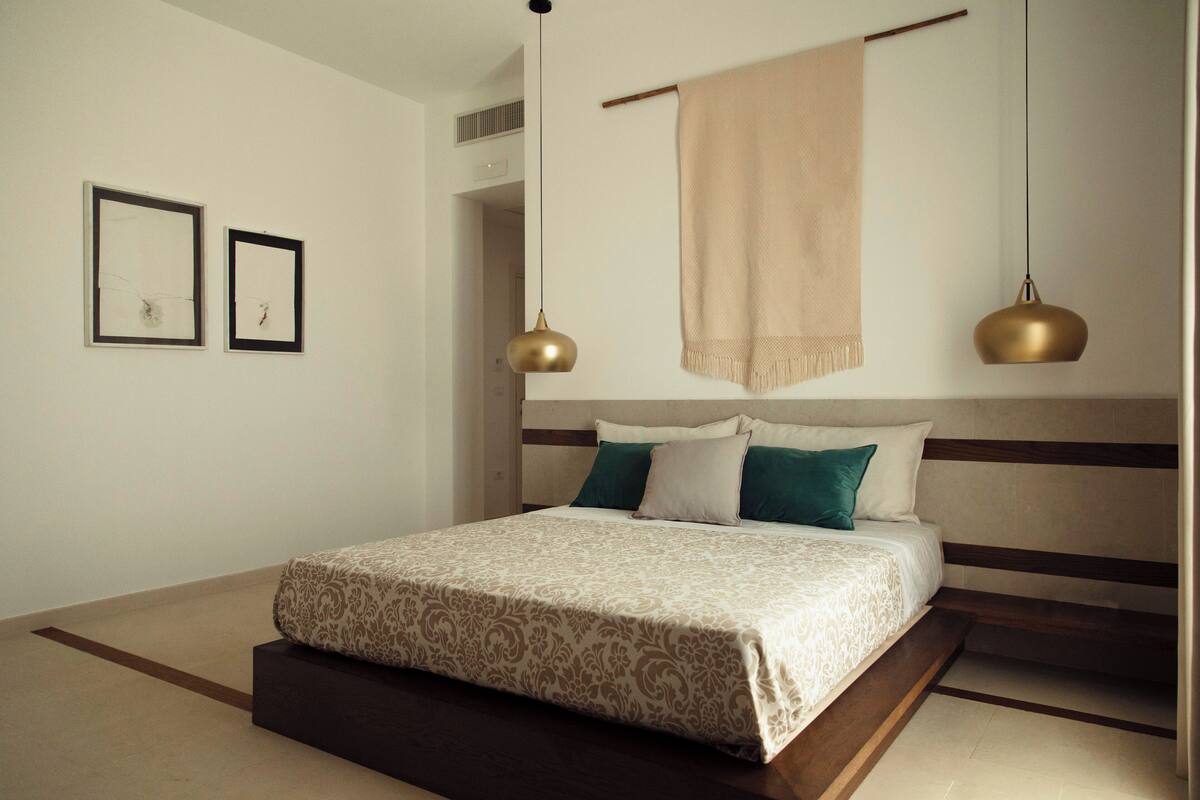 A modern bedroom with a large bed, two pendant lights, a headboard with horizontal stripes, and a beige throw blanket.