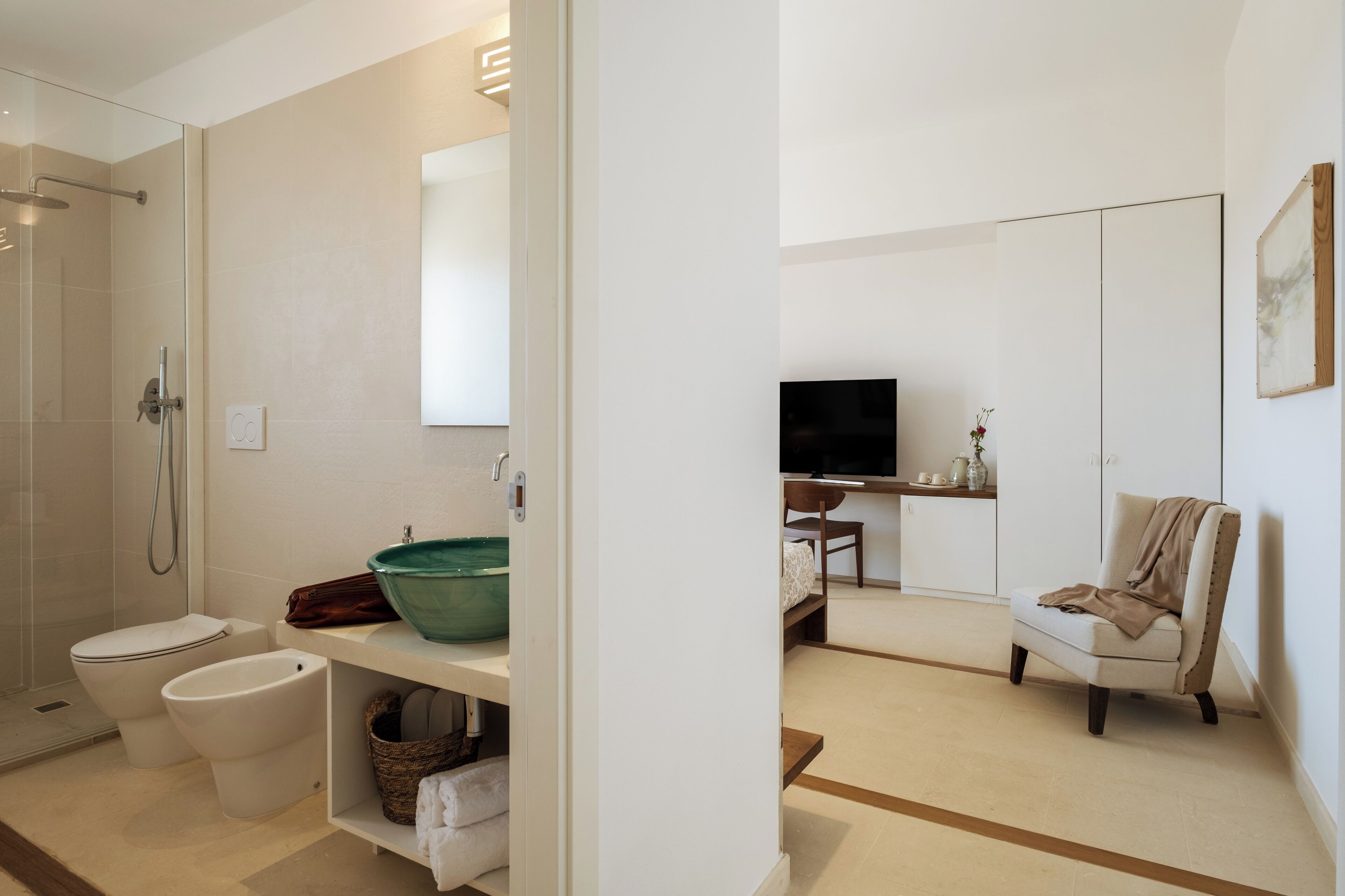 Un bagno moderno con doccia, toilette, bidet e lavandino con bacino verde. Nell'stanza adiacente c'è una sedia e un televisore.