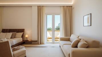 Quarto de hotel moderno com cama, sofá, poltrona, mesa de cabeceira com abajur e porta de vidro deslizante para área externa.