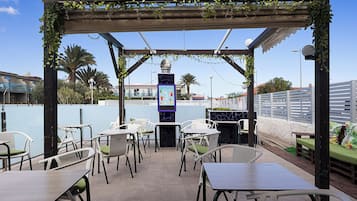 Um pátio ao ar livre com mesas e cadeiras, uma pérgula com vegetação e vista para o mar e edifícios ao fundo.