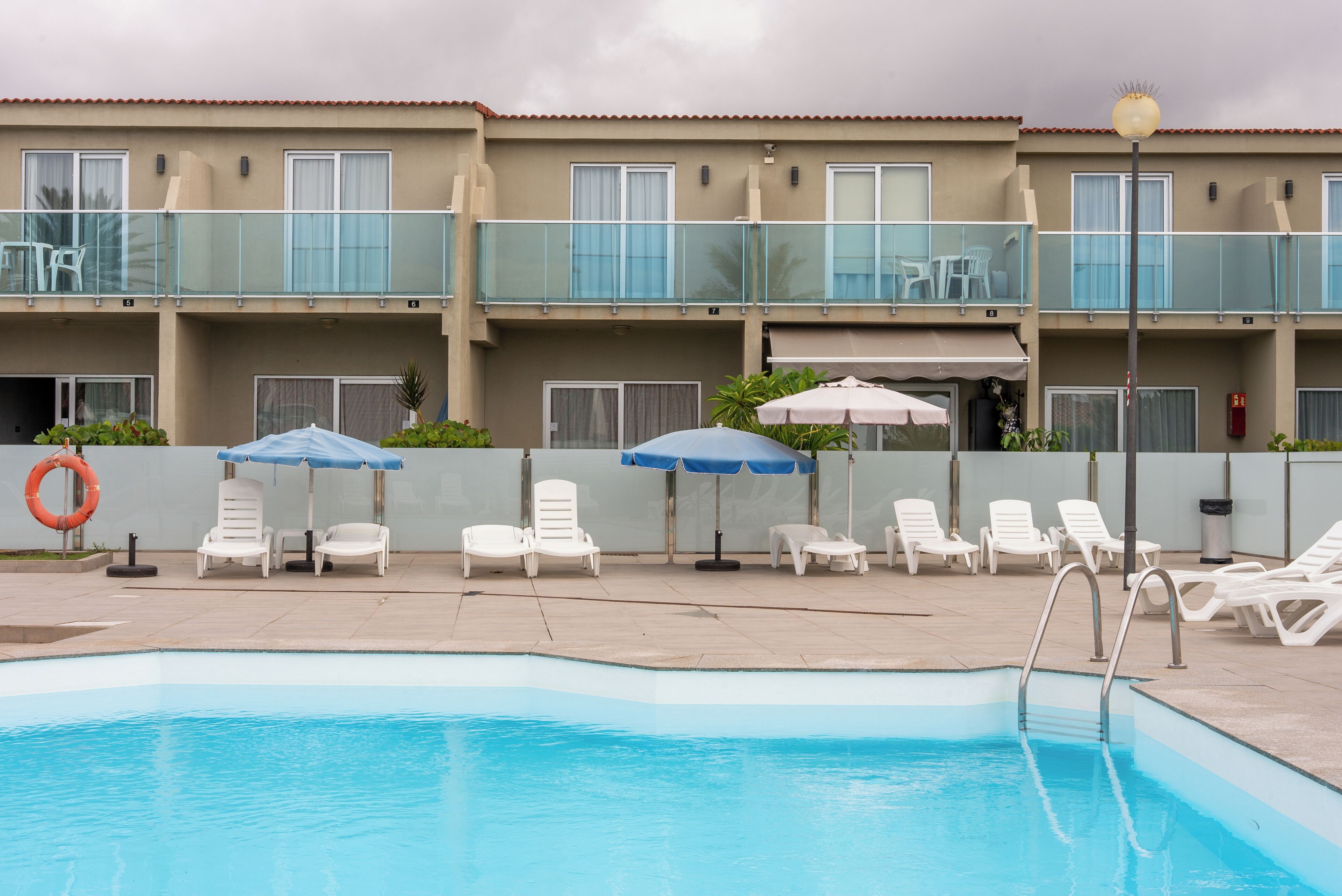 A swimming pool area with lounge chairs and umbrellas, a lifebuoy, and a multi-story building with balconies.