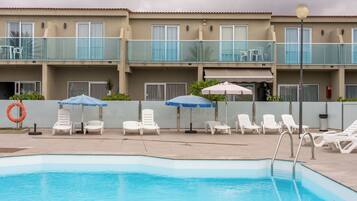A swimming pool area with lounge chairs and umbrellas, a lifebuoy, and a multi-story building with balconies.