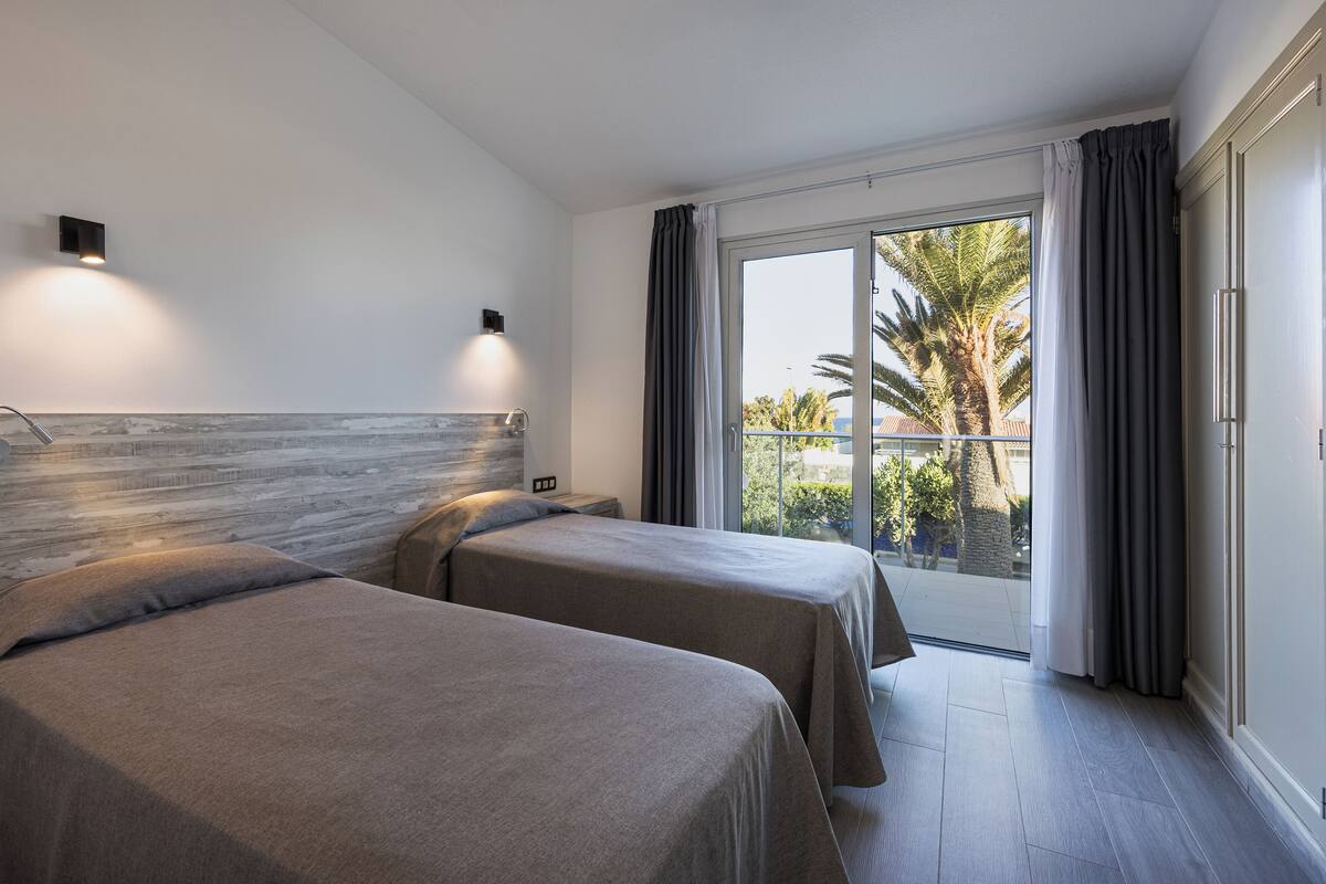 A hotel room with two beds, a wooden headboard, a nightstand, a wall-mounted lamp, and a view of palm trees and a balcony.