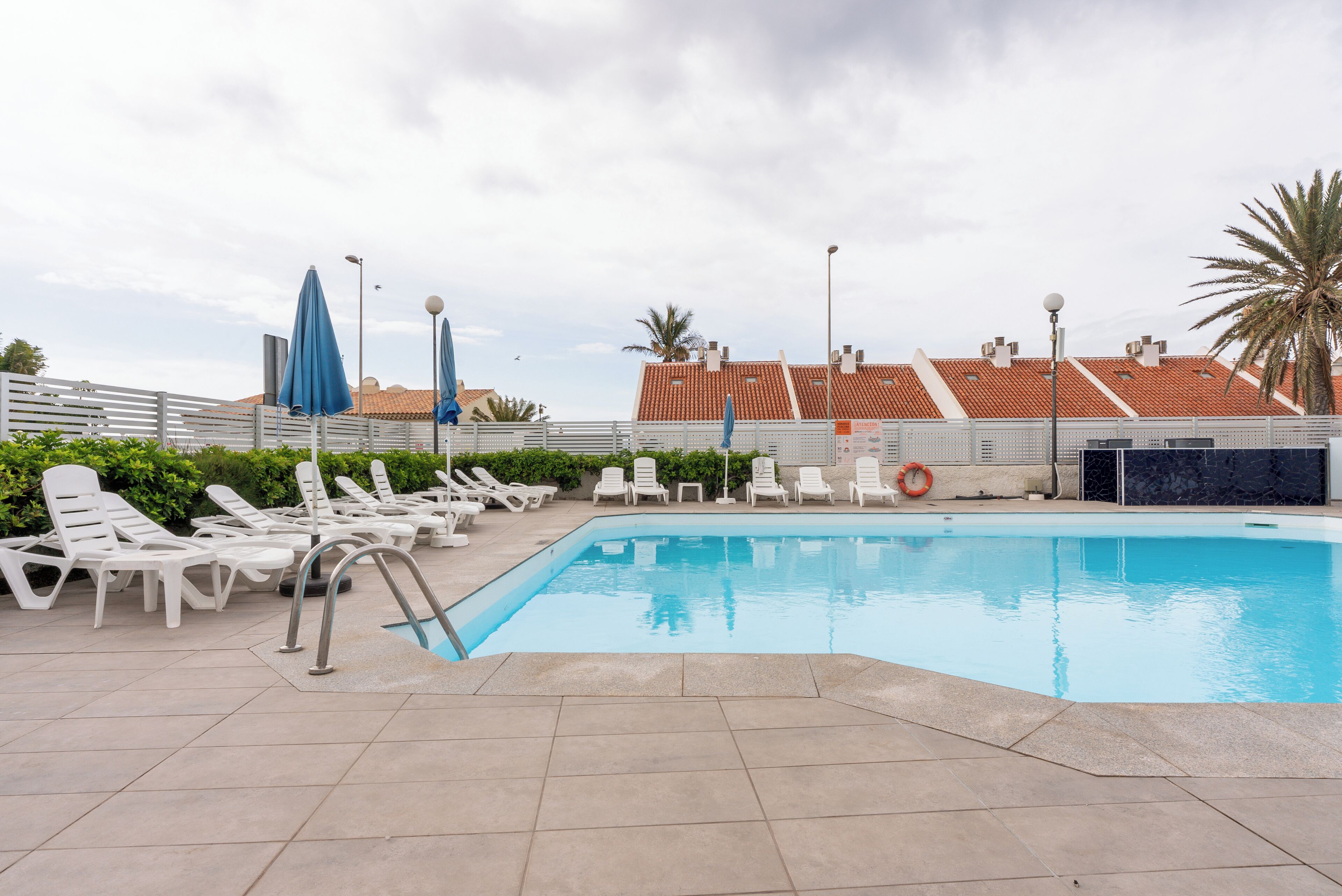 Una piscina con sillas de descanso y sombrillas, rodeada por una valla y palmeras.