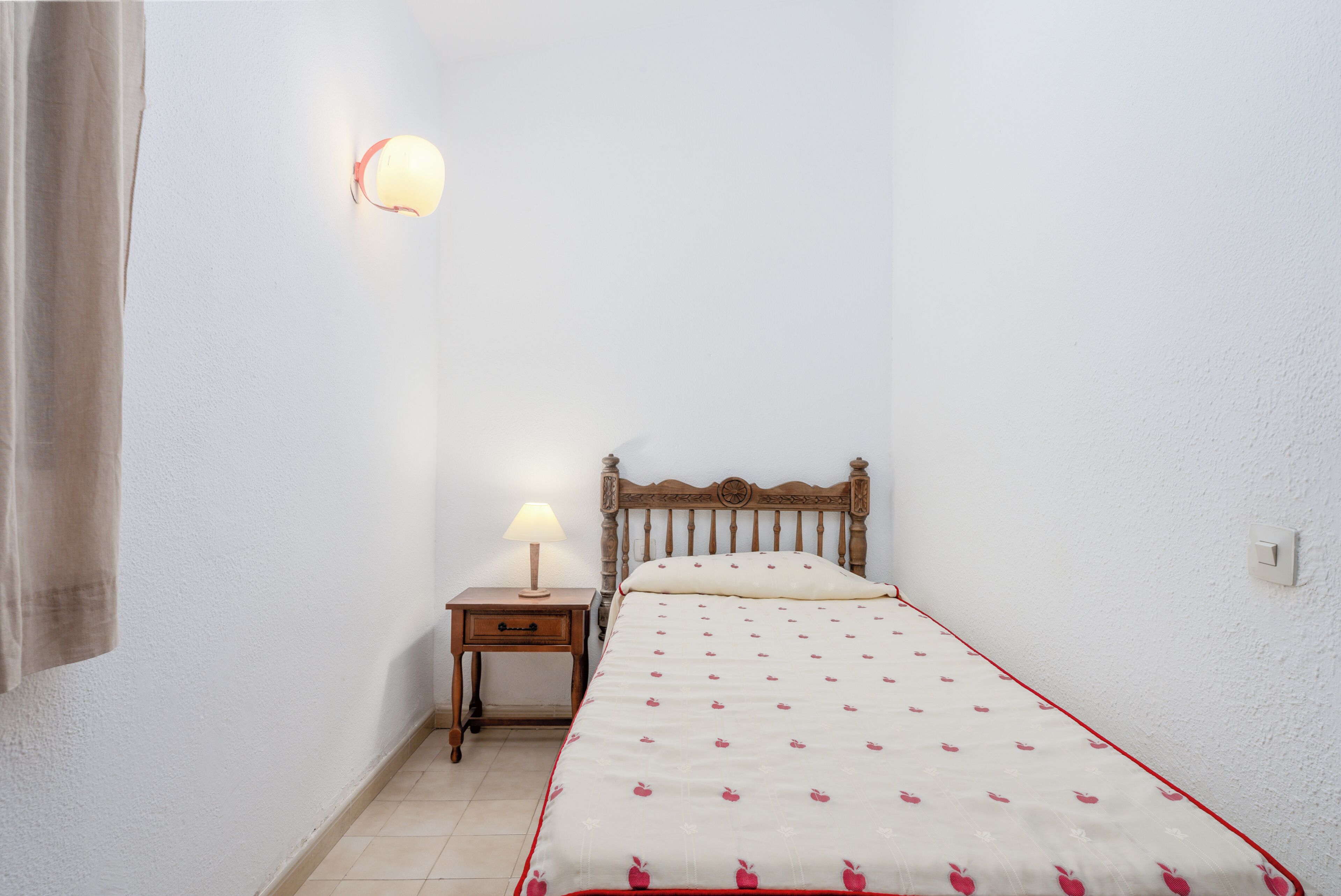 A small, neatly arranged bedroom with a single bed, a nightstand with a lamp, and a wall-mounted light fixture.