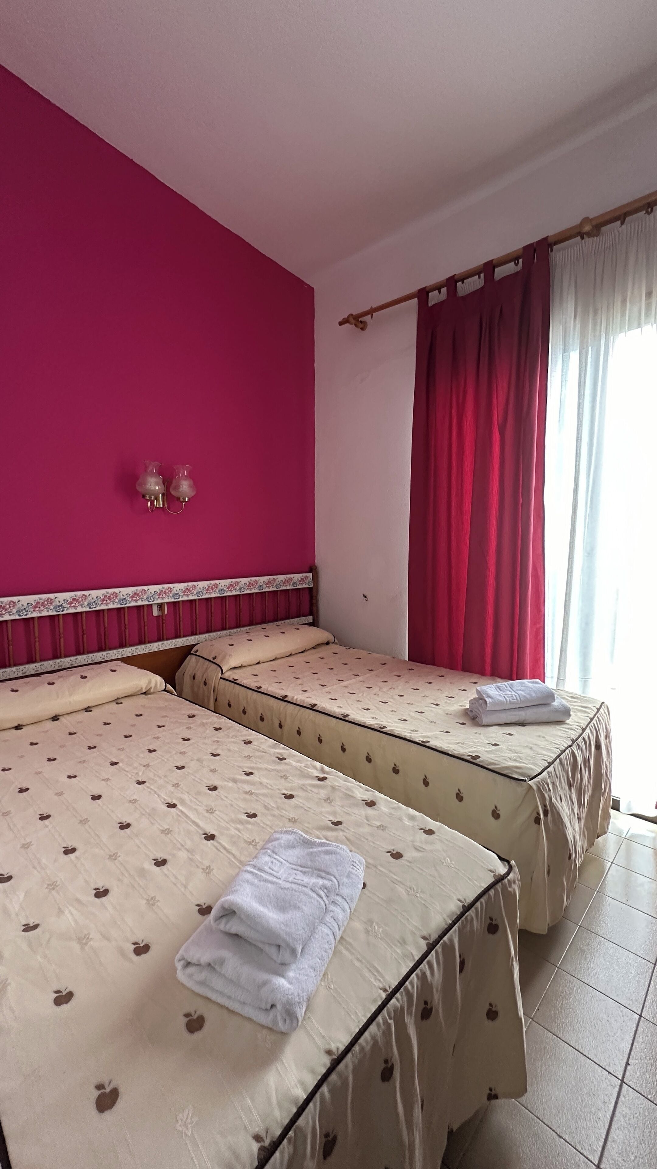 A hotel room with two beds, pink walls, and a window with red curtains.