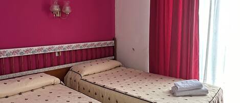 A hotel room with two beds, pink walls, and a window with red curtains.