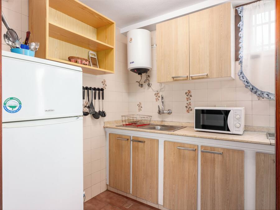 Eine kleine Küche mit Holzschränken, einem weißen Kühlschrank, einer Mikrowelle und einem Durchlauferhitzer.