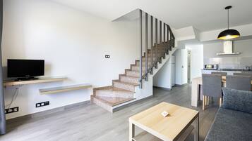 A modern living room with a TV on a glass shelf, a wooden coffee table, and a staircase leading to a kitchen area.
