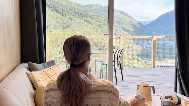 Een persoon zit op een bank, gericht op een balkon met uitzicht op bergen en een houten terras.