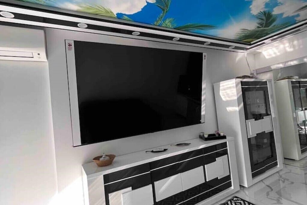 A modern living room with a large flat-screen TV mounted on a wall, a white and black entertainment unit below, and a ceiling mural depicting a tropical scene.