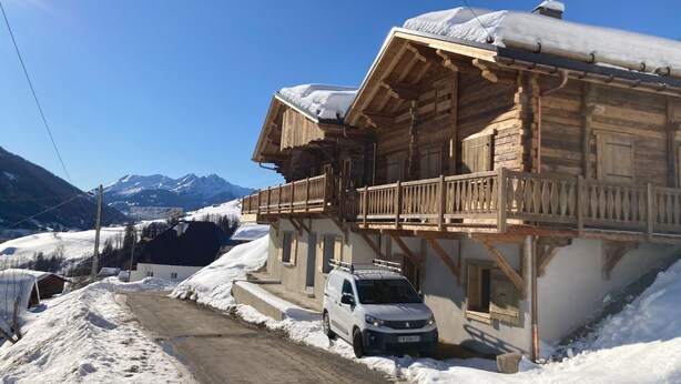 Una cabaña de madera con balcón, un coche estacionado al frente y montañas nevadas al fondo.