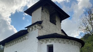 Un edificio rústico blanco con techo de madera y un veleta con forma de gallo, rodeado de follaje y una cerca de madera.