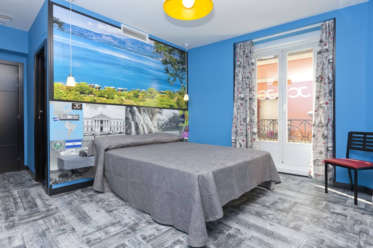 A bedroom with a bed, a window with curtains, a chair, and a wall with a mural.