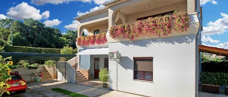 A two-story house with a balcony adorned with flowers, a staircase leading to the upper floor, and a red car parked in the driveway.
