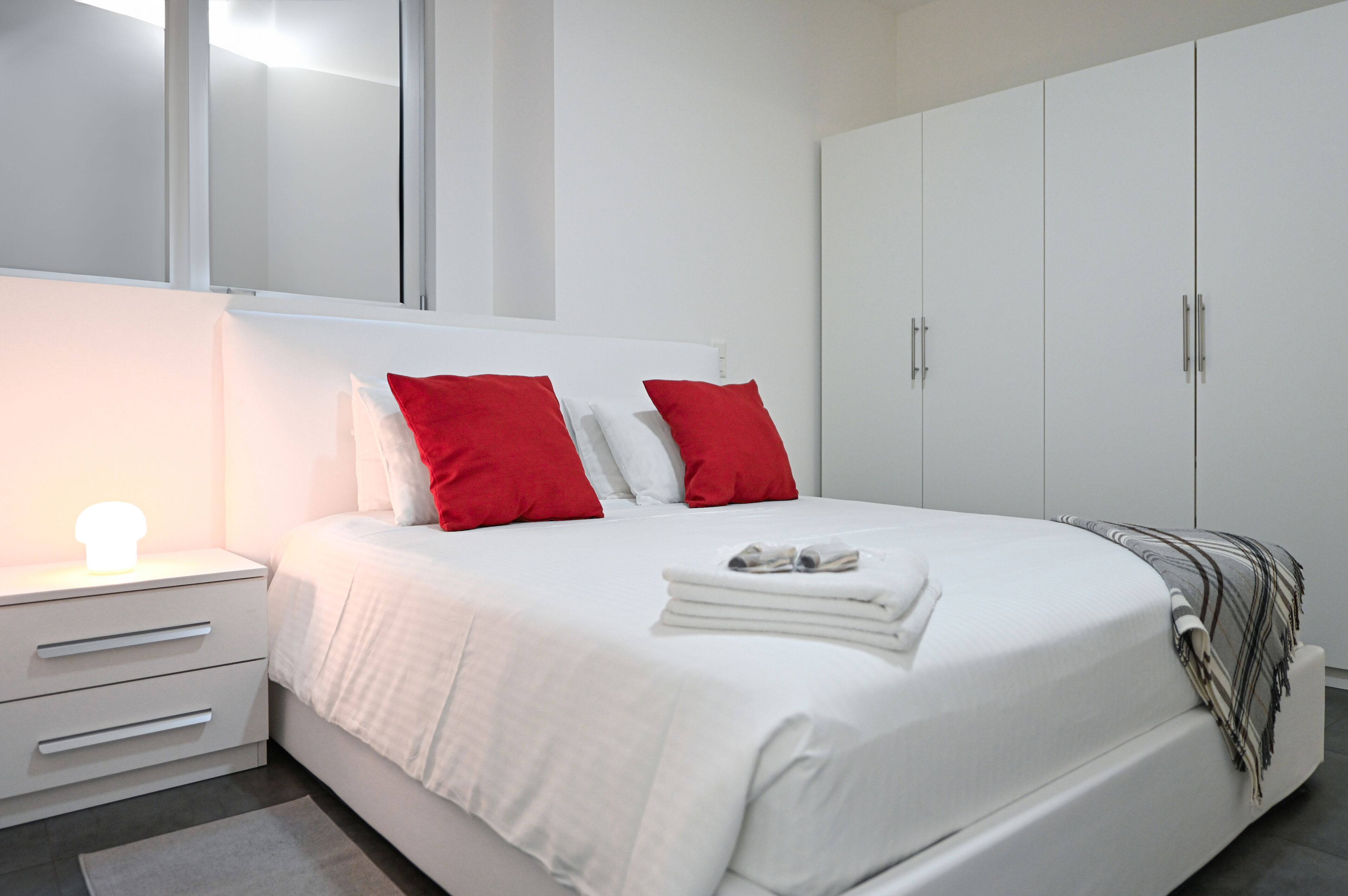 A modern bedroom with a large bed, white bedding, red pillows, a bedside lamp, and a wardrobe.