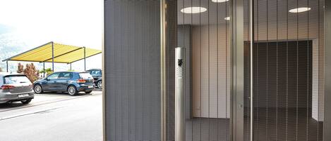 Entrada de un edificio con una puerta de metal, una zona de espera techada y vista a coches estacionados.