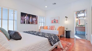 Un dormitorio con cama, sofá, un cuadro en la pared, lámpara y baño visible a través de una puerta abierta.