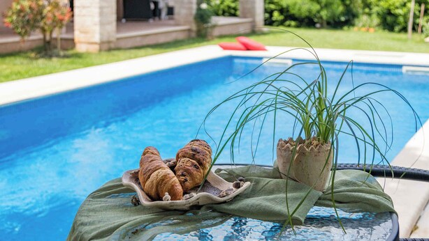 A poolside setting with a table holding croissants and a potted plant. - Holiday apartment with pool (Ližnjan)