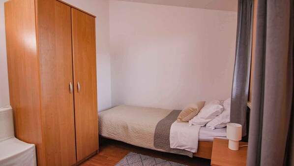 Una habitación pequeña con una cama individual, un armario de madera, una mesita de noche con lámpara y una alfombra gris.