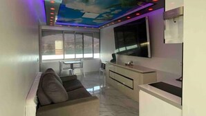 Un espace de vie compact comprenant un canapé, une table à manger, un coin cuisine et un téléviseur fixé au mur.