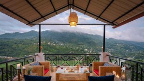 Un balcón con mobiliario de mimbre, una mesa con vasos y aperitivos, y vistas a montañas verdes.