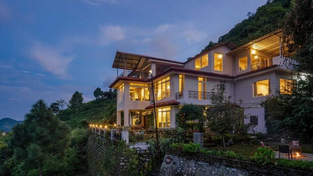 Una casa de dos pisos con varios balcones, rodeada de abundante vegetación y un muro de piedra.