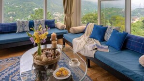 Una habitación con un sofá frente a un amplio ventanal con vistas a un paisaje montañoso. Hay una mesa de centro de vidrio con una cesta de aperitivos y una vela.