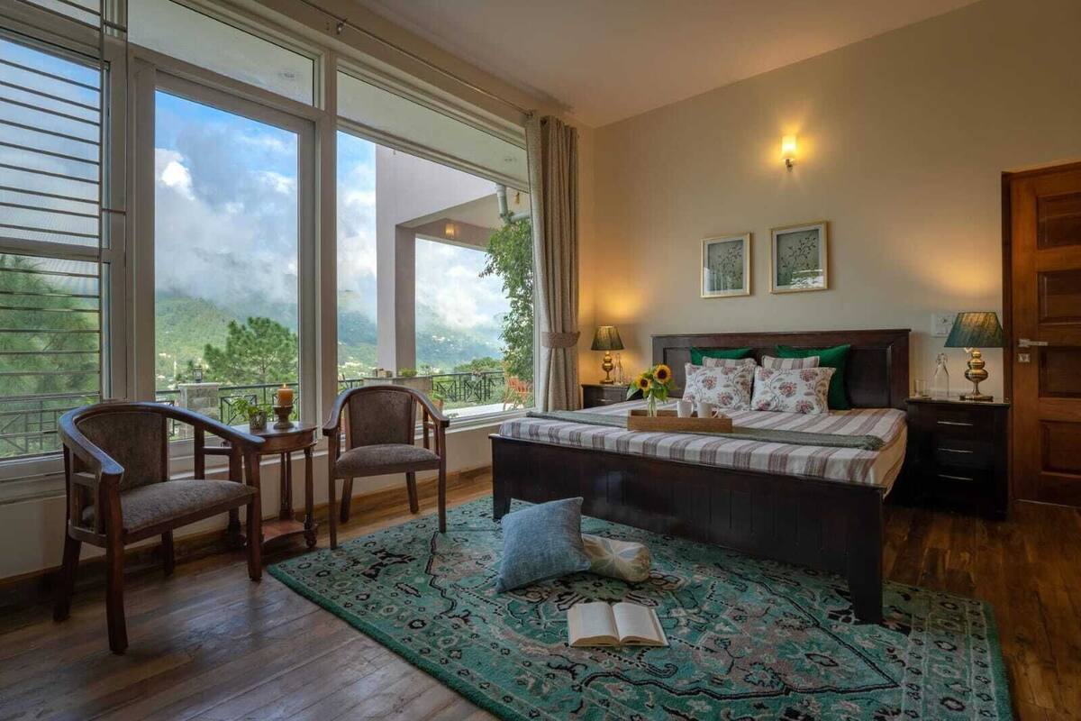 A bedroom with a large window, a bed with floral bedding, two chairs, a bedside table with a lamp, and a view of the outdoors.