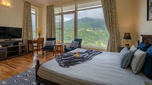 Habitación de hotel con una cama grande, un escritorio con televisor, dos sillas, vistas a un paisaje montañoso a través de amplias ventanas y una alfombra estampada.