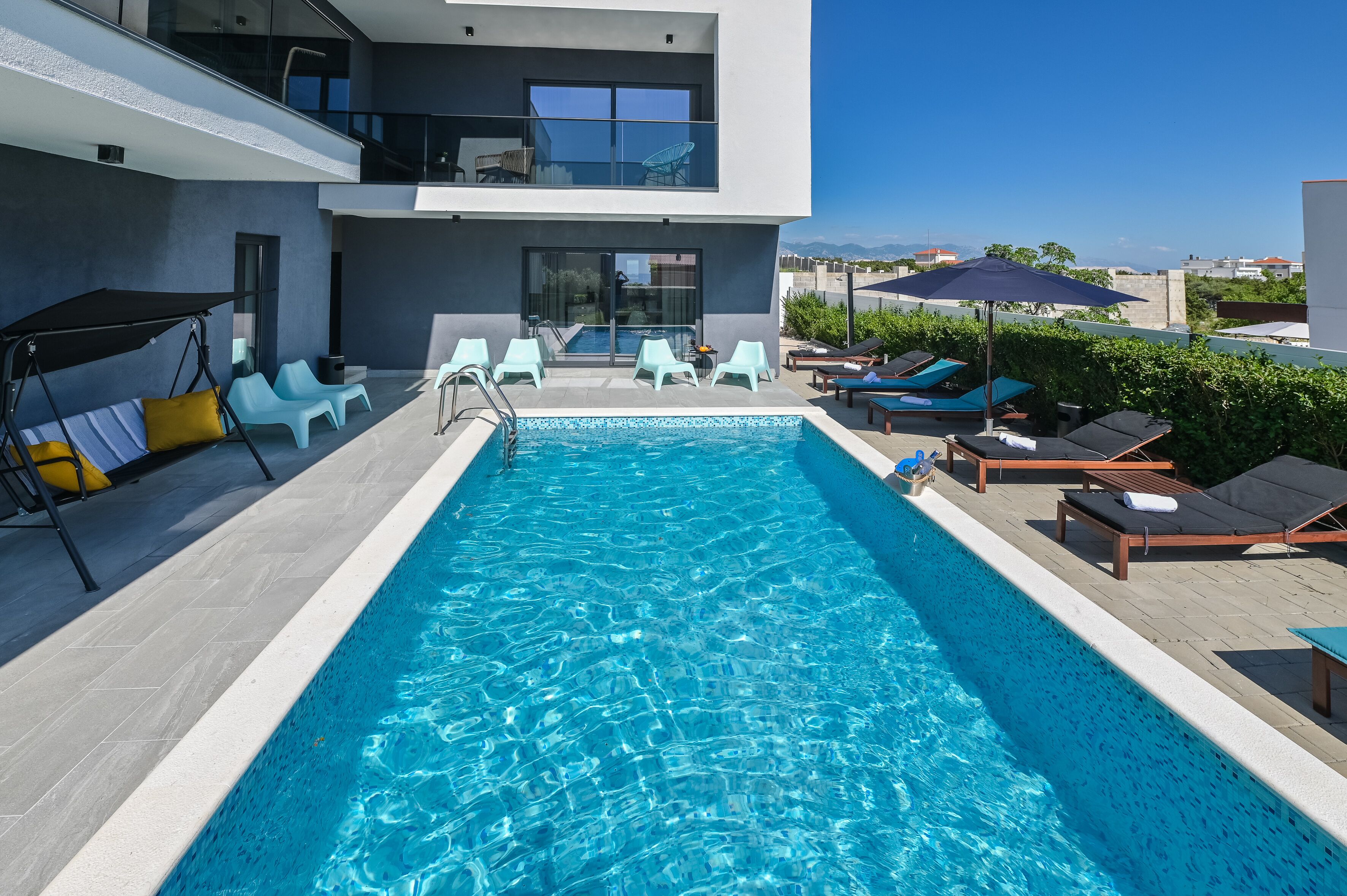 Uma piscina moderna ao ar livre com espreguiçadeiras e uma cadeira de balanço, cercada por um edifício com varanda de vidro.